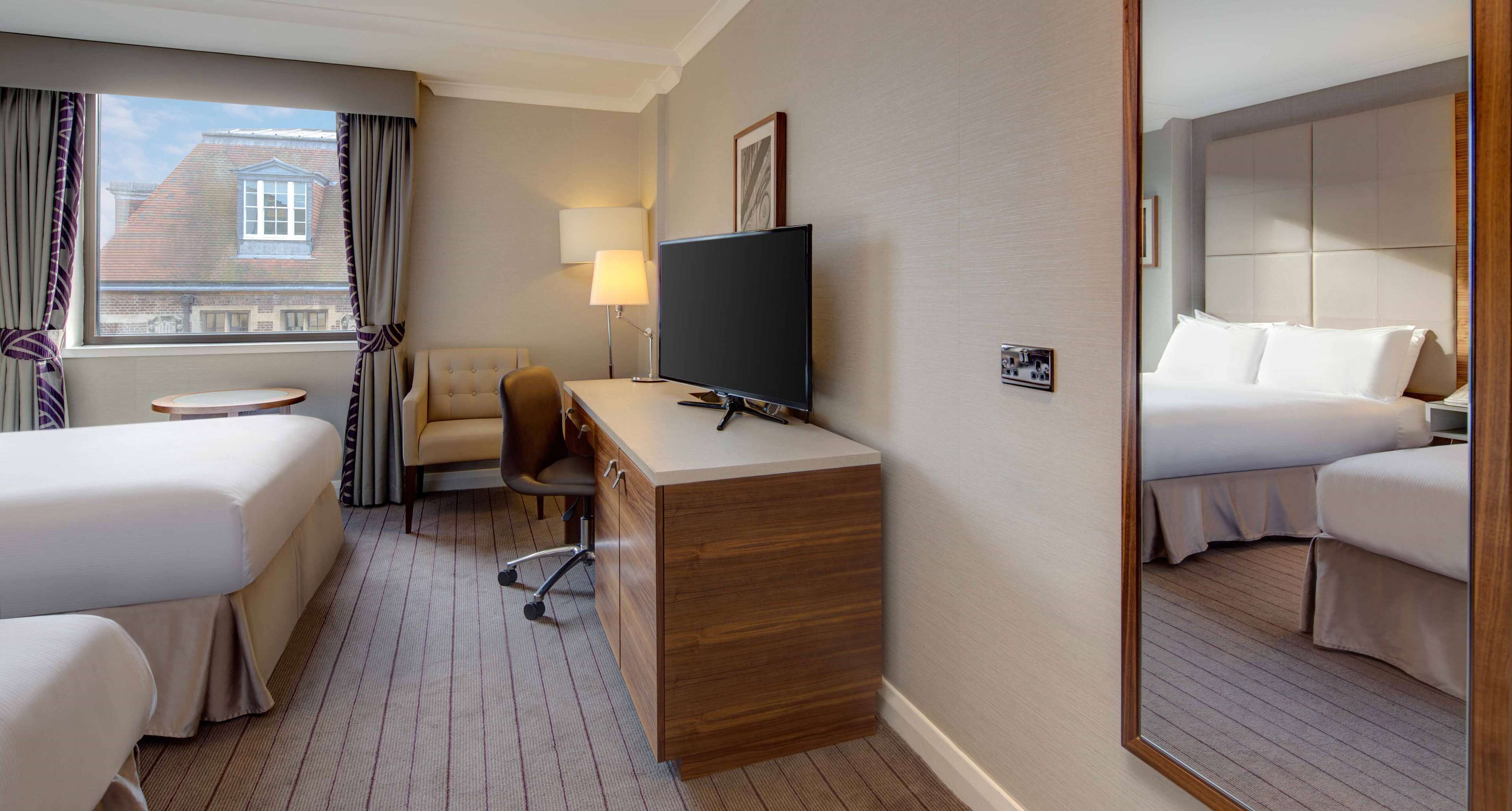 A hotel room with two beds, a desk with a flat-screen TV, a chair, a lamp, and a window with curtains.