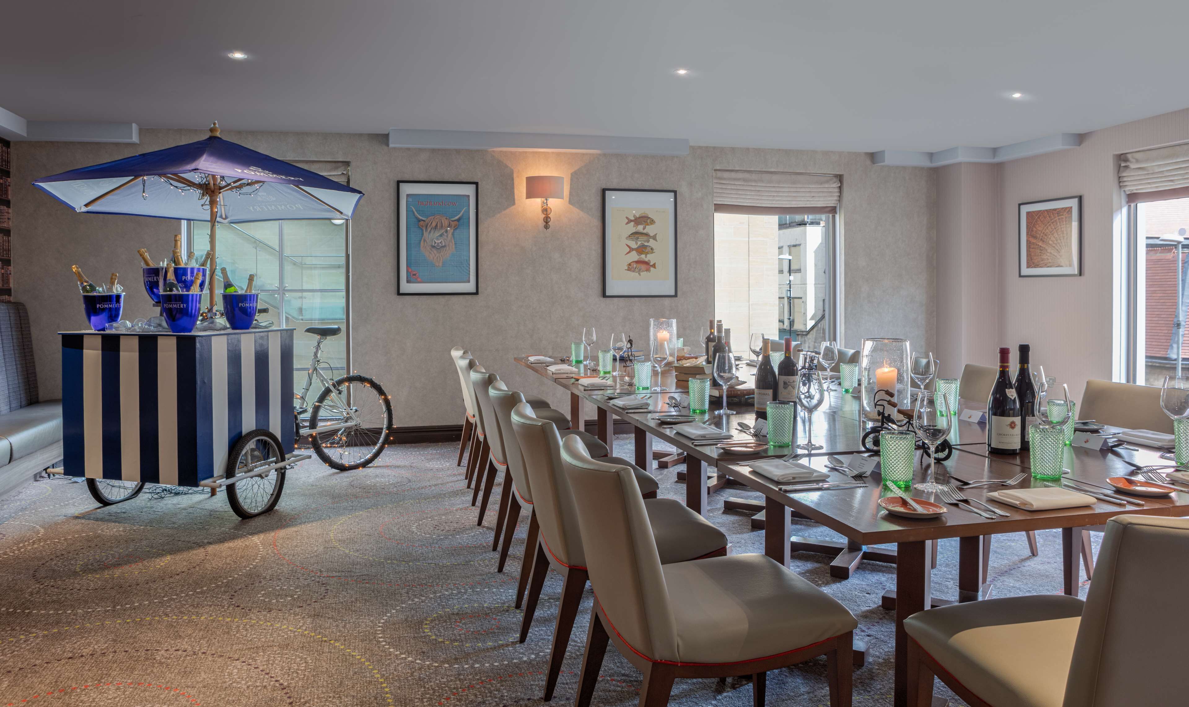 A dining area with a long table set for six, surrounded by chairs. There is a bicycle with a blue umbrella and champagne buckets on the left. The room has a carpeted floor and a large window with a view.