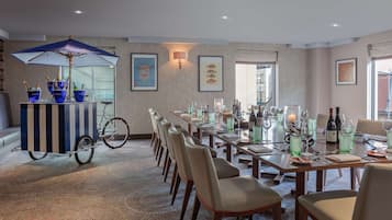 Un comedor con una mesa larga dispuesta para seis personas, rodeada de sillas. A la izquierda, una bicicleta con un paraguas azul y cubos de champán. La habitación tiene suelo alfombrado y un ventanal con vistas.