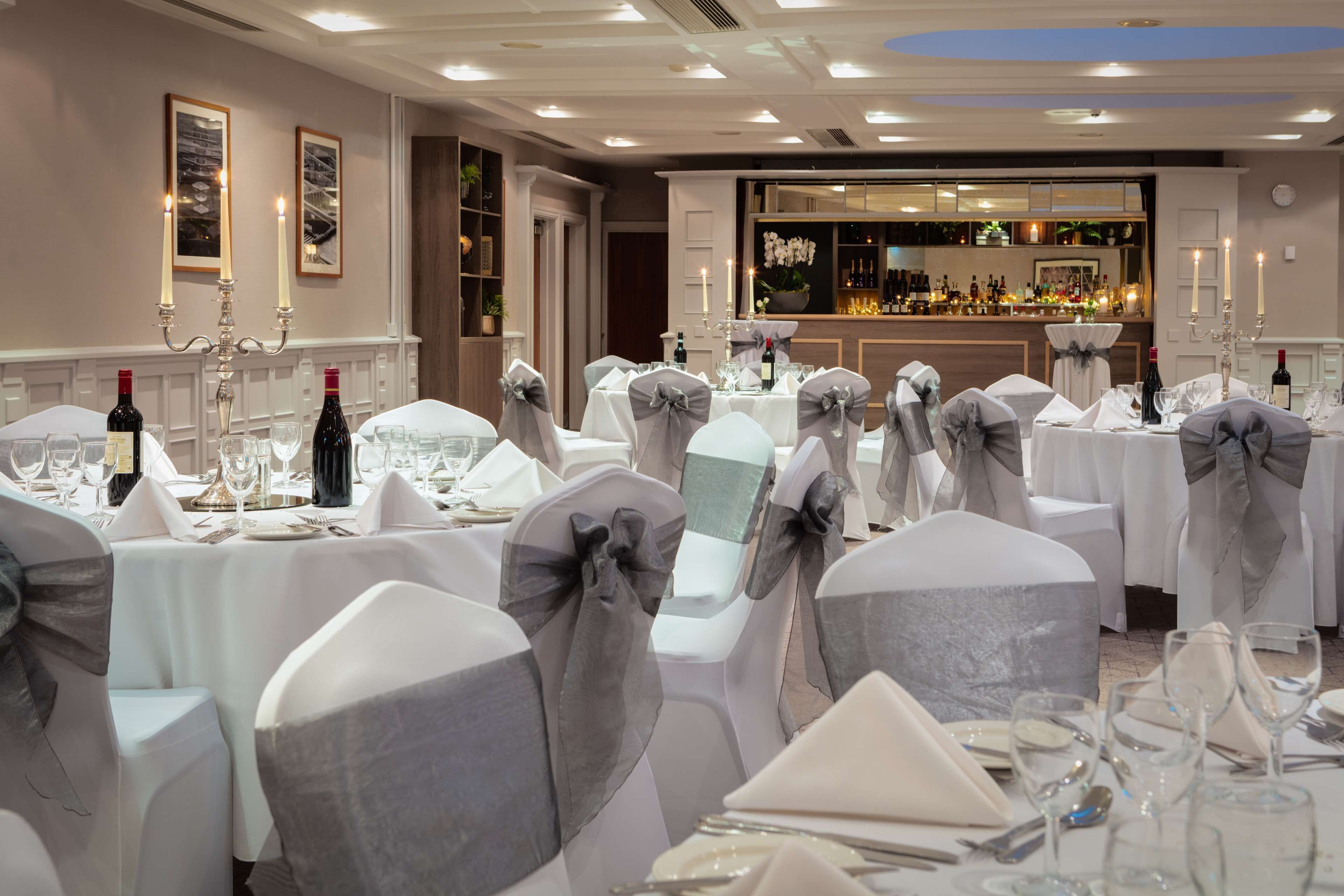 A banquet hall with round tables set for a formal event, featuring white tablecloths, grey chair covers, and candelabras.