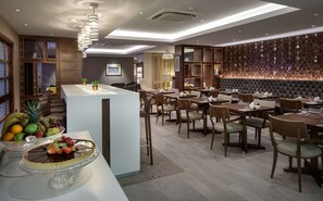 A modern restaurant with a central bar area, multiple dining tables, and a fruit display. - Hilton Cambridge City Centre (Cambridge)