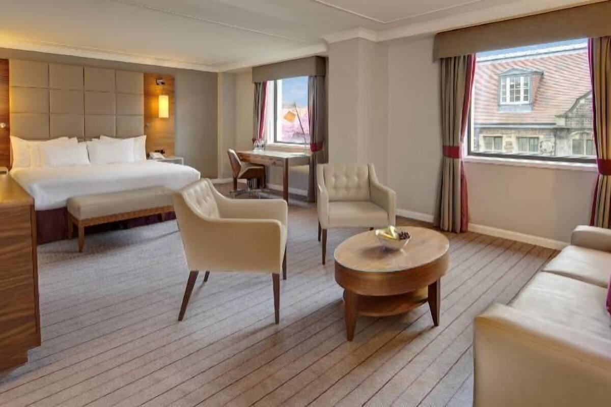 Habitación de hotel con cama, dos sillones, un sofá, una mesa de centro de madera, un escritorio y una ventana con cortinas.