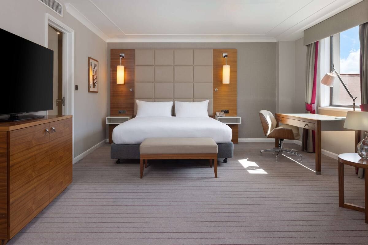 Una habitación de hotel moderna con una cama grande, mesitas de noche, un escritorio, una silla, un televisor y una ventana con cortinas.