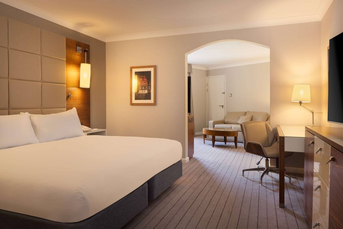 Habitación de hotel con una cama grande, un escritorio, una silla y un sofá. Se observa un cuadro en la pared y una lámpara sobre el escritorio.
