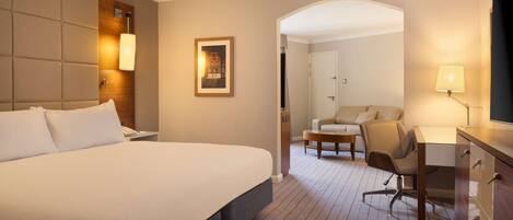 A hotel room with a large bed, a desk, a chair, and a sofa. There is a framed picture on the wall and a lamp on the desk.