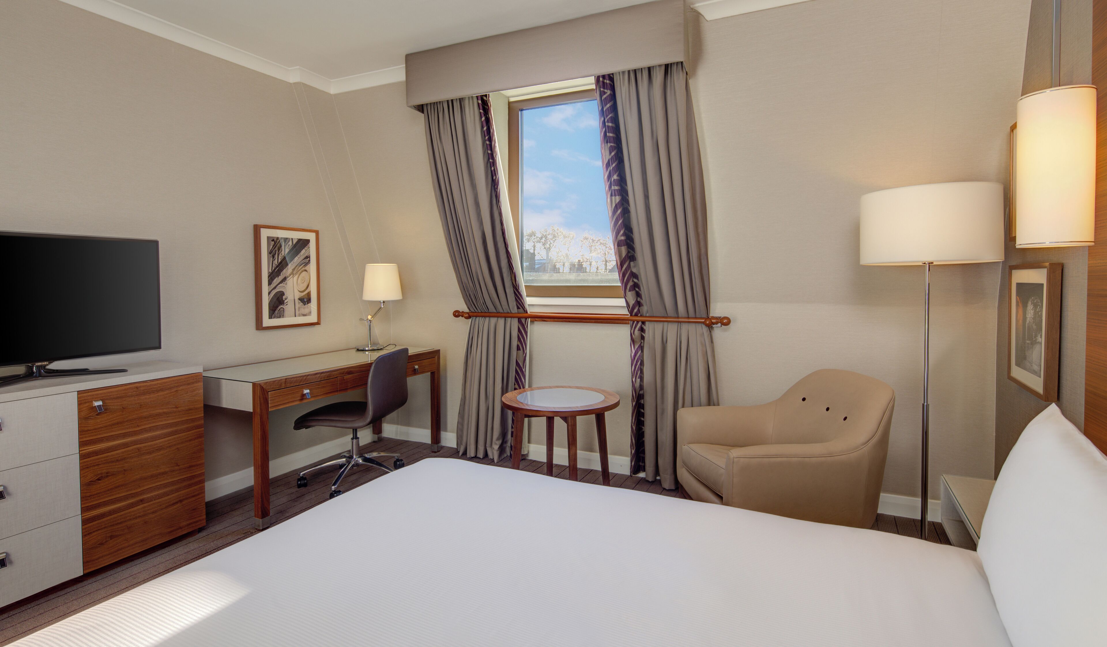 Quarto de hotel com cama, escrivaninha, cadeira, televisão e janela com cortinas.
