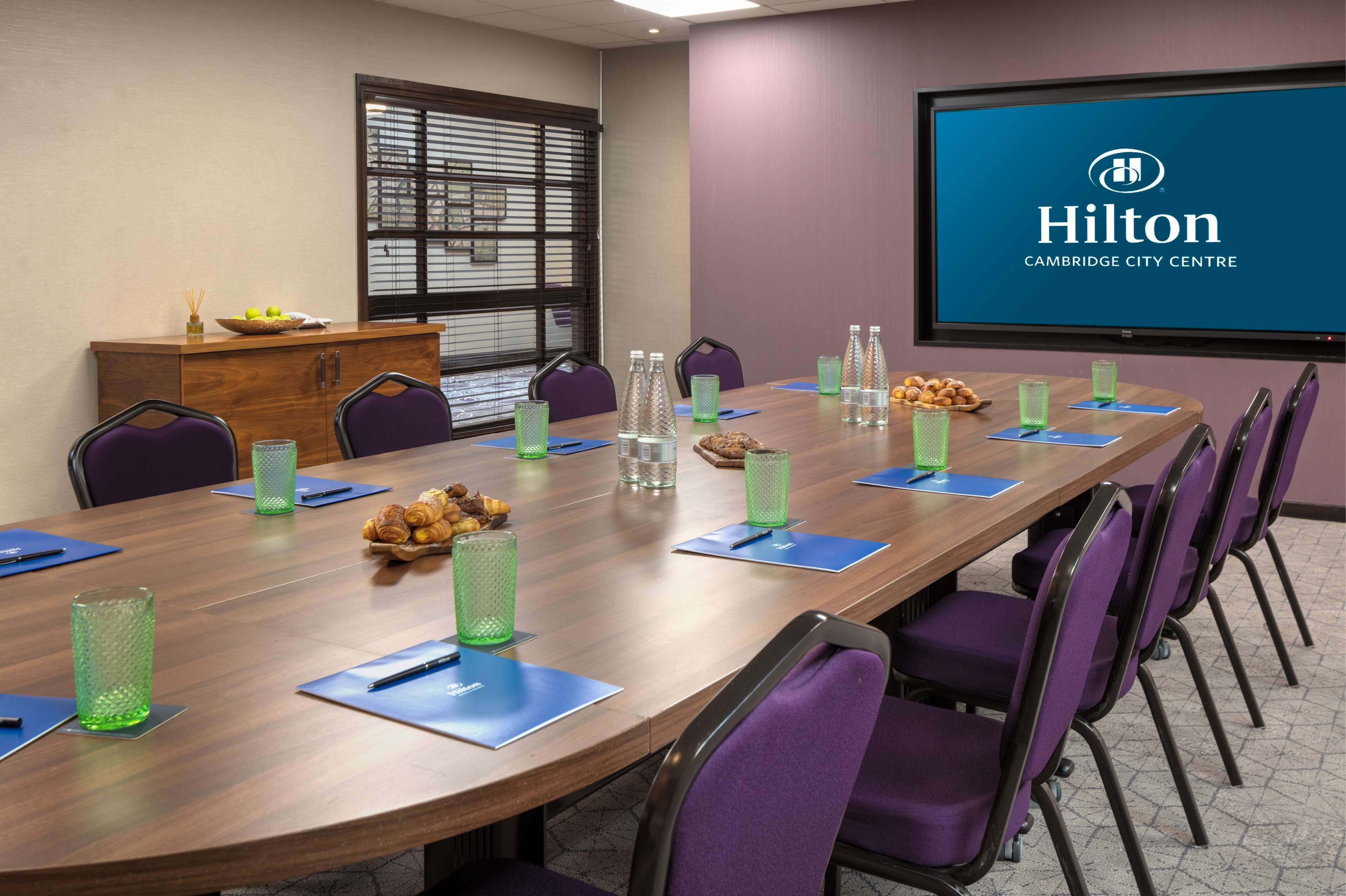 A conference room with a long table, purple chairs, and a projector screen displaying 'Hilton Cambridge City Centre'.
