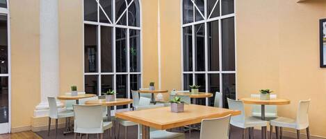 A modern indoor dining area with round tables and white chairs, large arched windows, and a wooden floor.