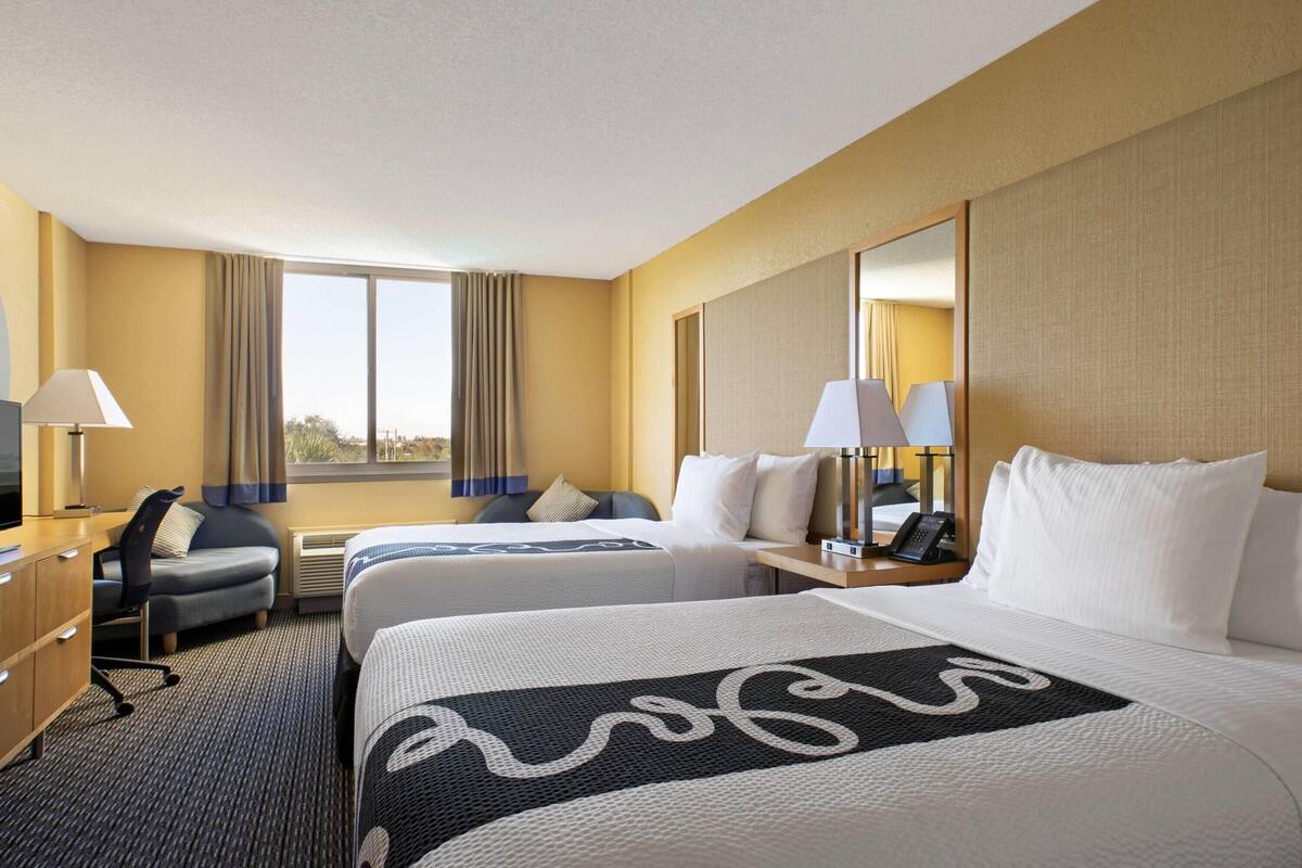 Ein Hotelzimmer mit zwei Betten, einem Schreibtisch, einem Stuhl, einer Lampe und einem großen Fenster.