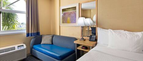 A hotel room with a bed, a sofa, a desk with a lamp, a phone, and a window with curtains.