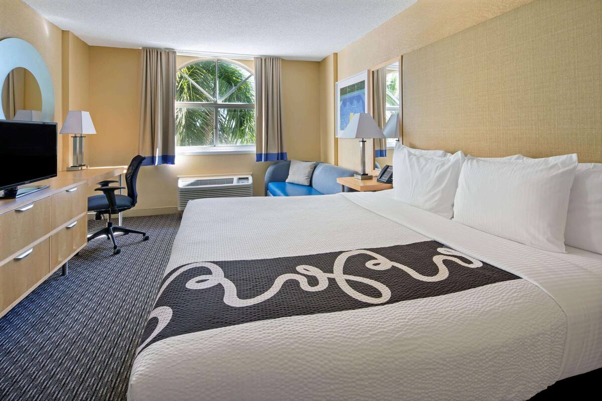 Ein Hotelzimmer mit einem großen Bett, einem Schreibtisch mit Fernseher, einem Sessel und einem Fenster mit Vorhängen.