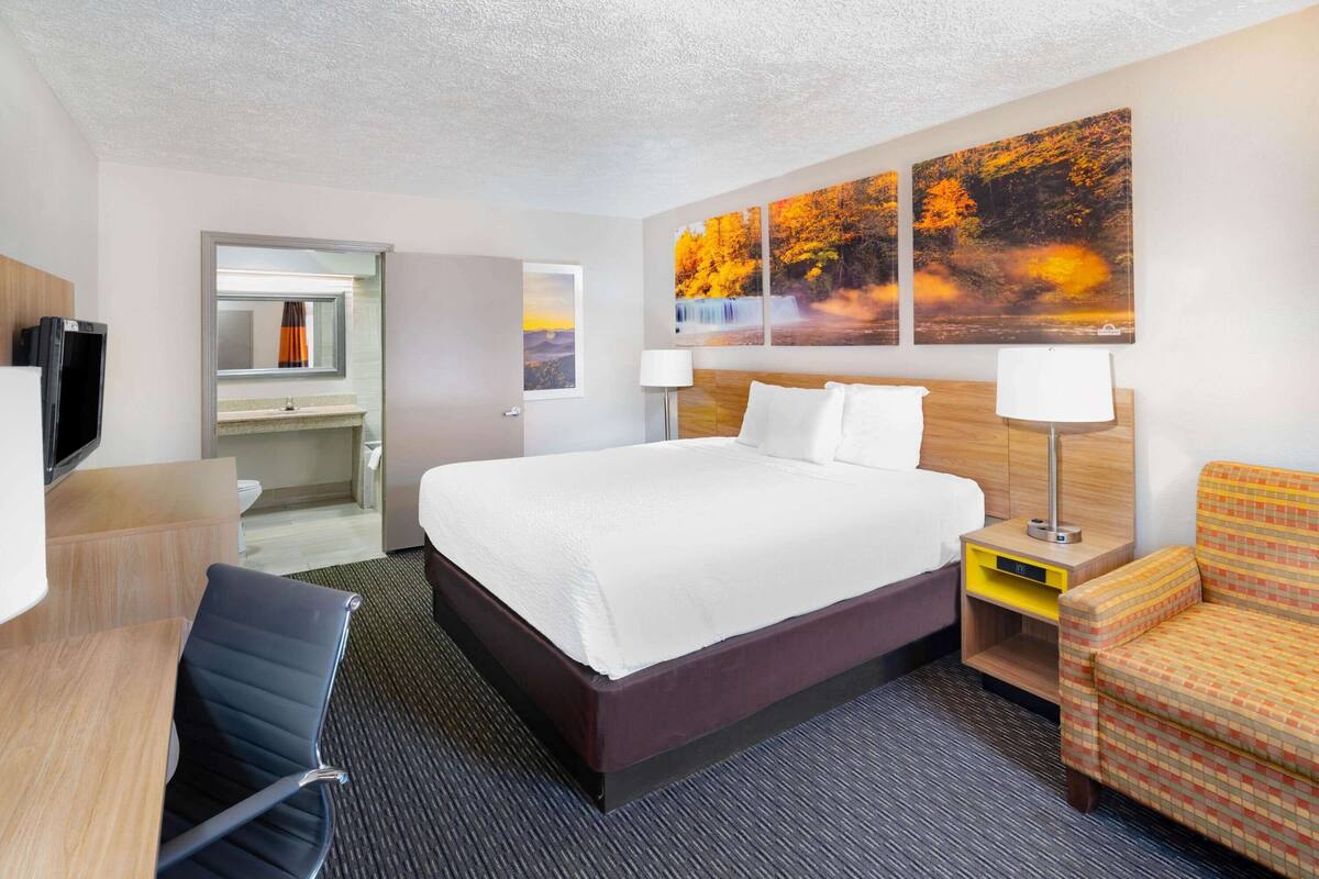 Ein Hotelzimmer mit einem großen Bett, einem Schreibtisch mit Stuhl, einem Fernseher und einem Badezimmer, sowie einer Wand mit herbstlichen Bildern.