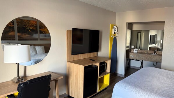 Una habitación de hotel con cama, televisión, escritorio con lámpara, espejo y una pequeña kitchenette.