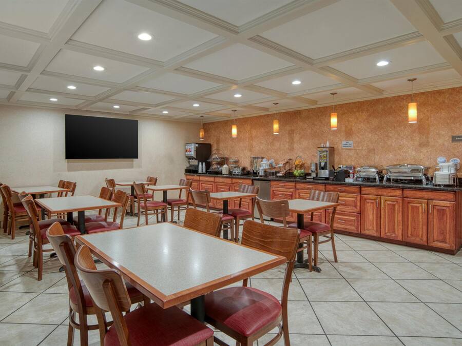 A dining area with multiple tables and chairs, a kitchen counter with various food items, and a large screen on the wall.