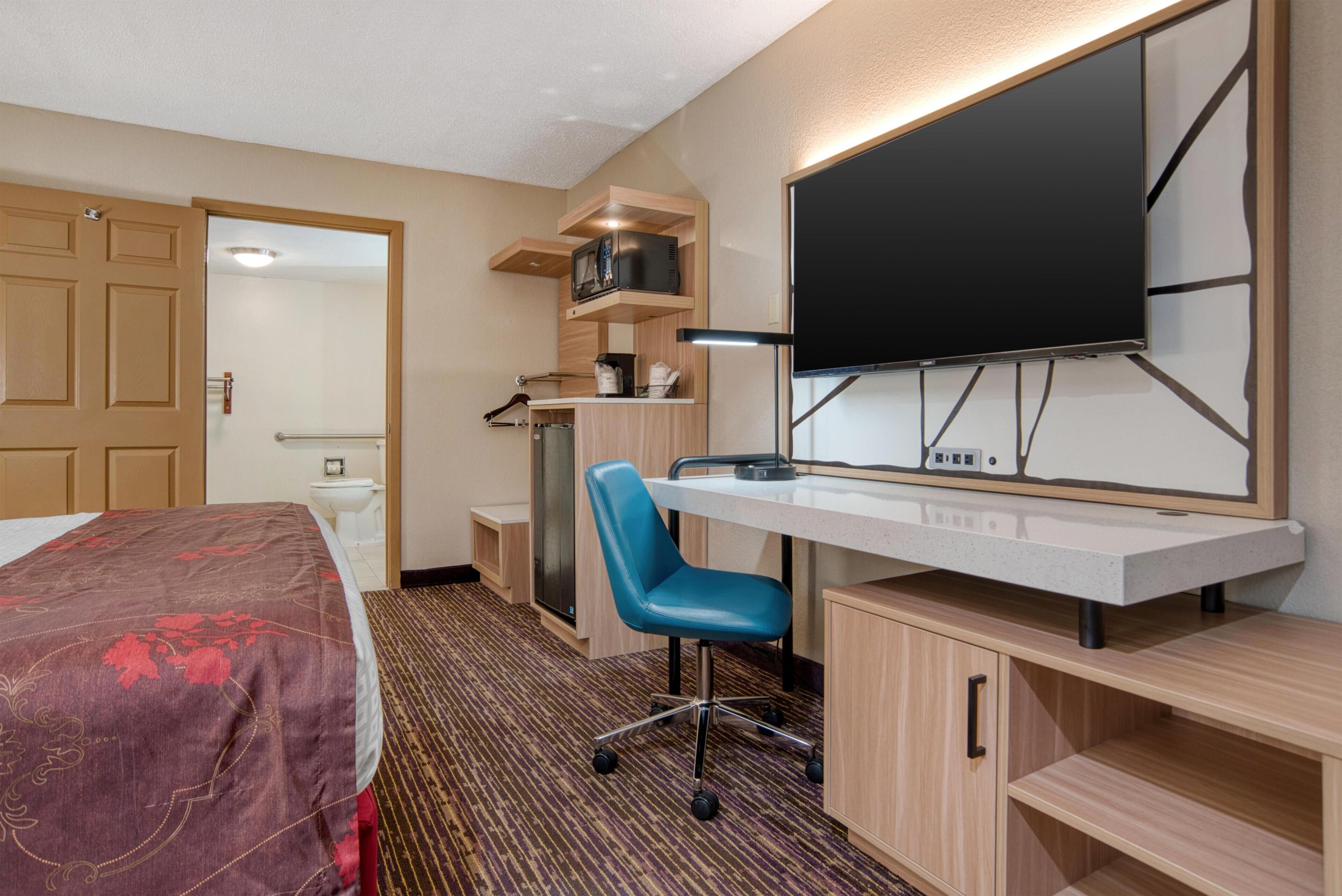 A hotel room with a bed, desk, chair, TV, microwave, and a small kitchenette.