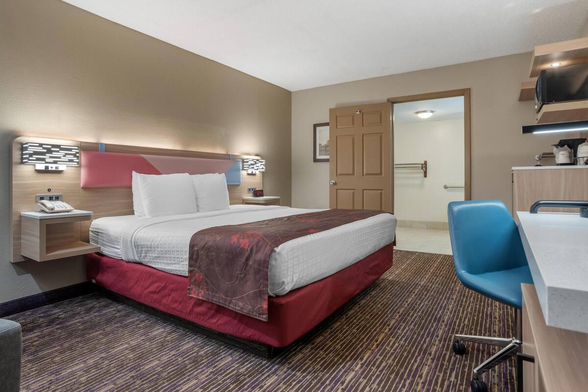 Een hotelkamer met een bed, nachtkastje, lamp, bureau, stoel en een deur naar een badkamer.