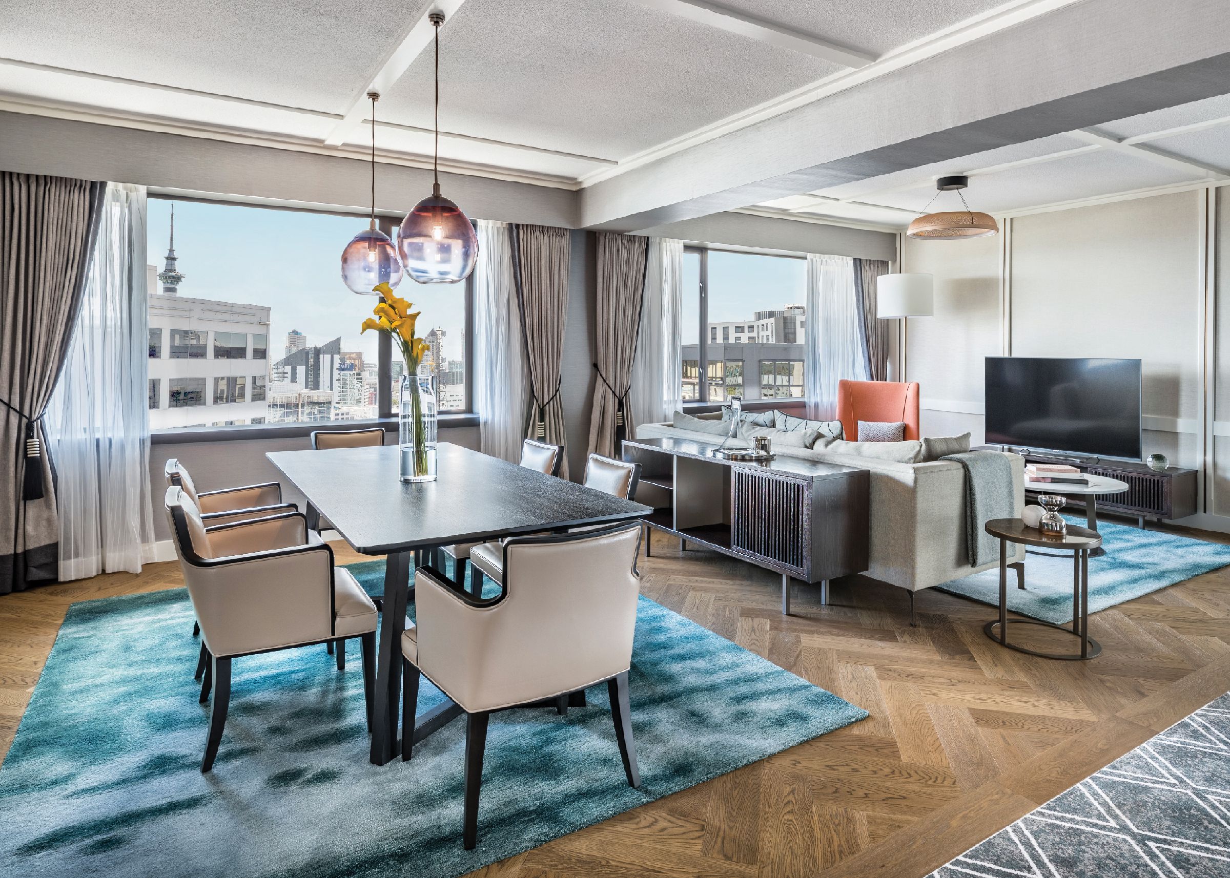 A modern hotel room with a dining area, a sofa, a TV, and a view of the cityscape.