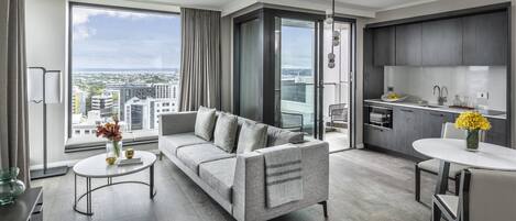 A modern living room with a grey sofa, a round coffee table, and a dining area with a white table and chairs. A large window offers a view of the cityscape.