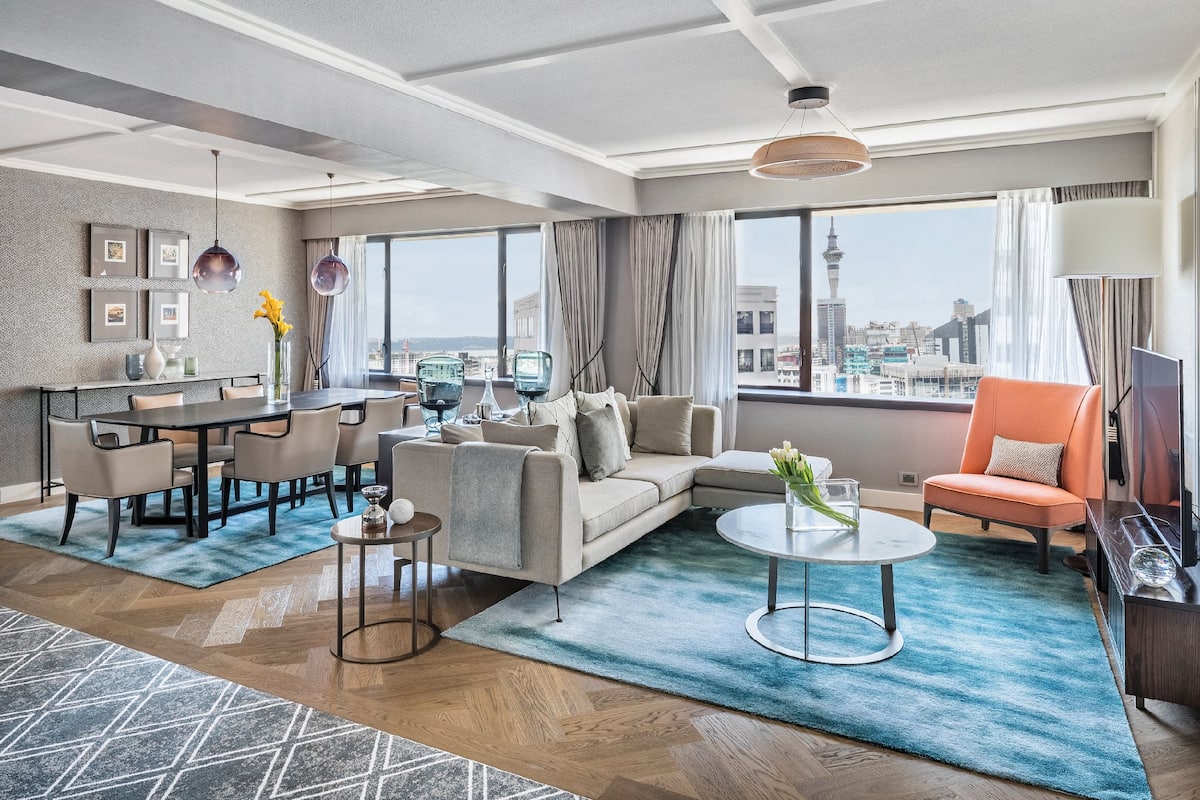 A modern hotel room with a dining area, a sofa, a coffee table, and a view of a cityscape.
