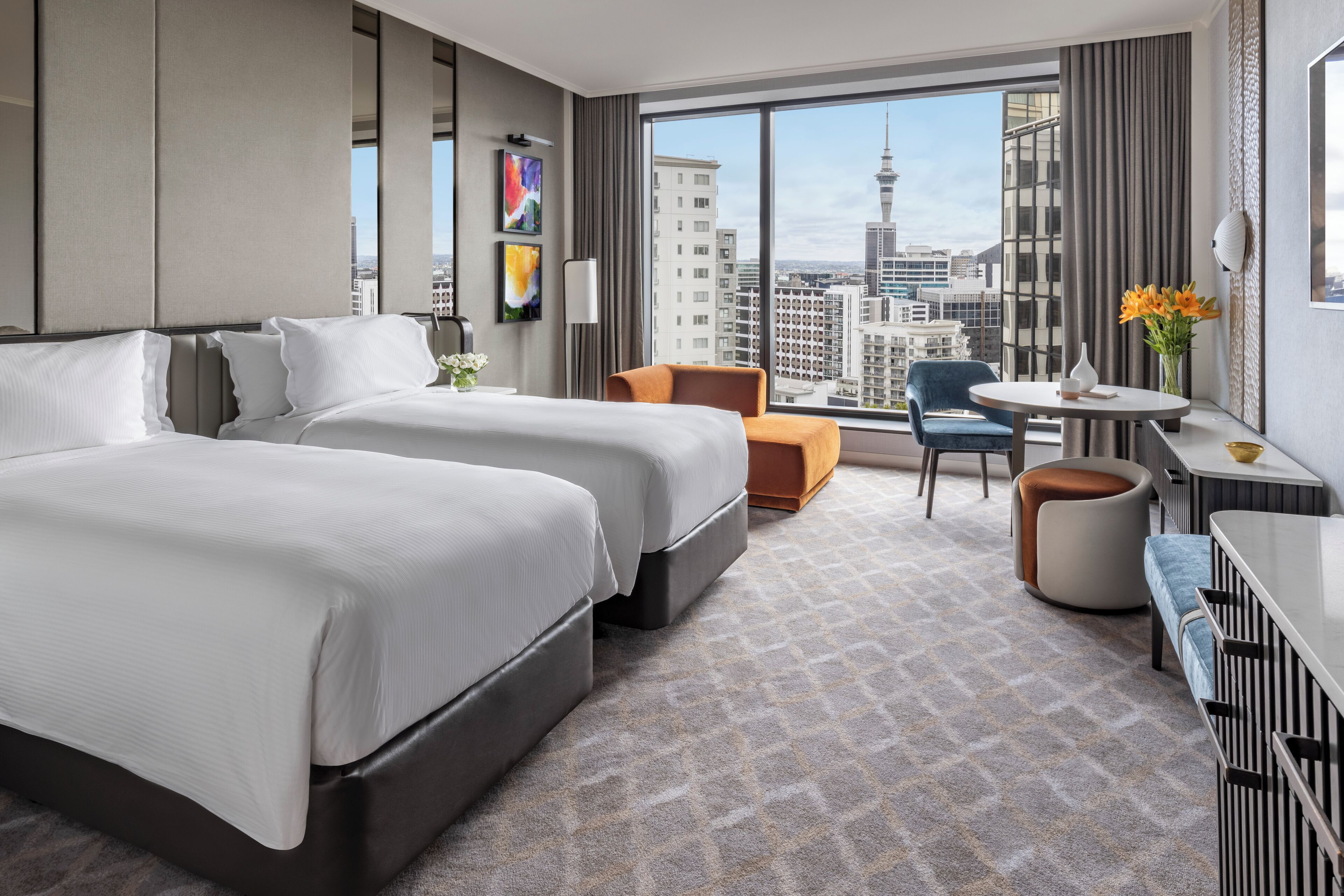 A hotel room with two beds, a seating area with a table and chairs, and a large window offering a city view.