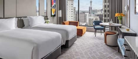 A hotel room with two beds, a seating area with a table and chairs, and a large window offering a city view.