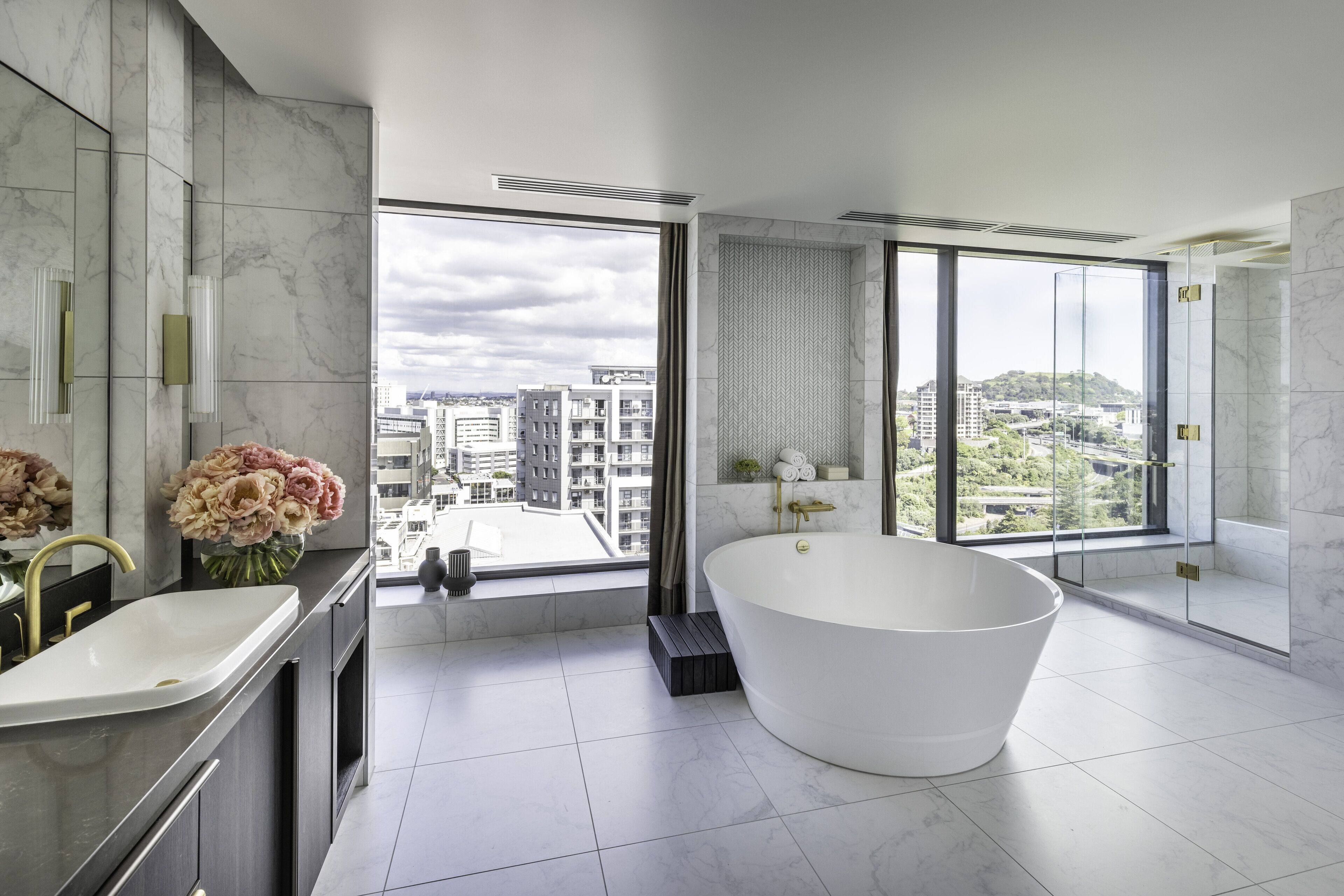 Un baño moderno con una bañera independiente, un amplio ventanal con vista a la ciudad y un tocador de mármol.