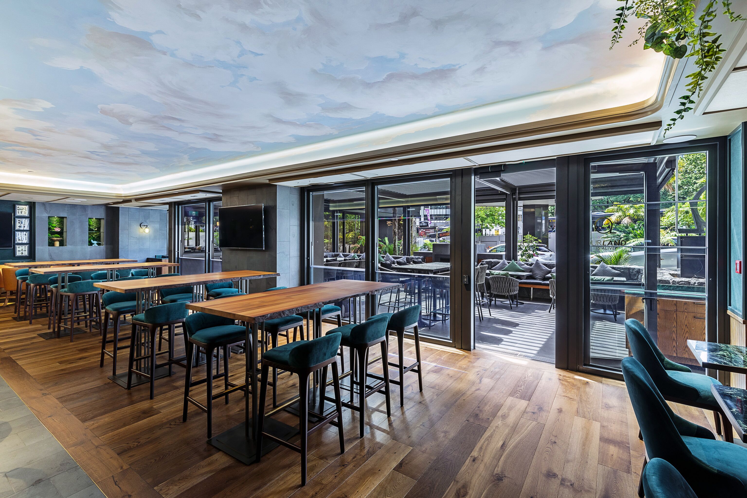 A modern indoor dining area with wooden tables and teal chairs, large windows, and a ceiling mural.
