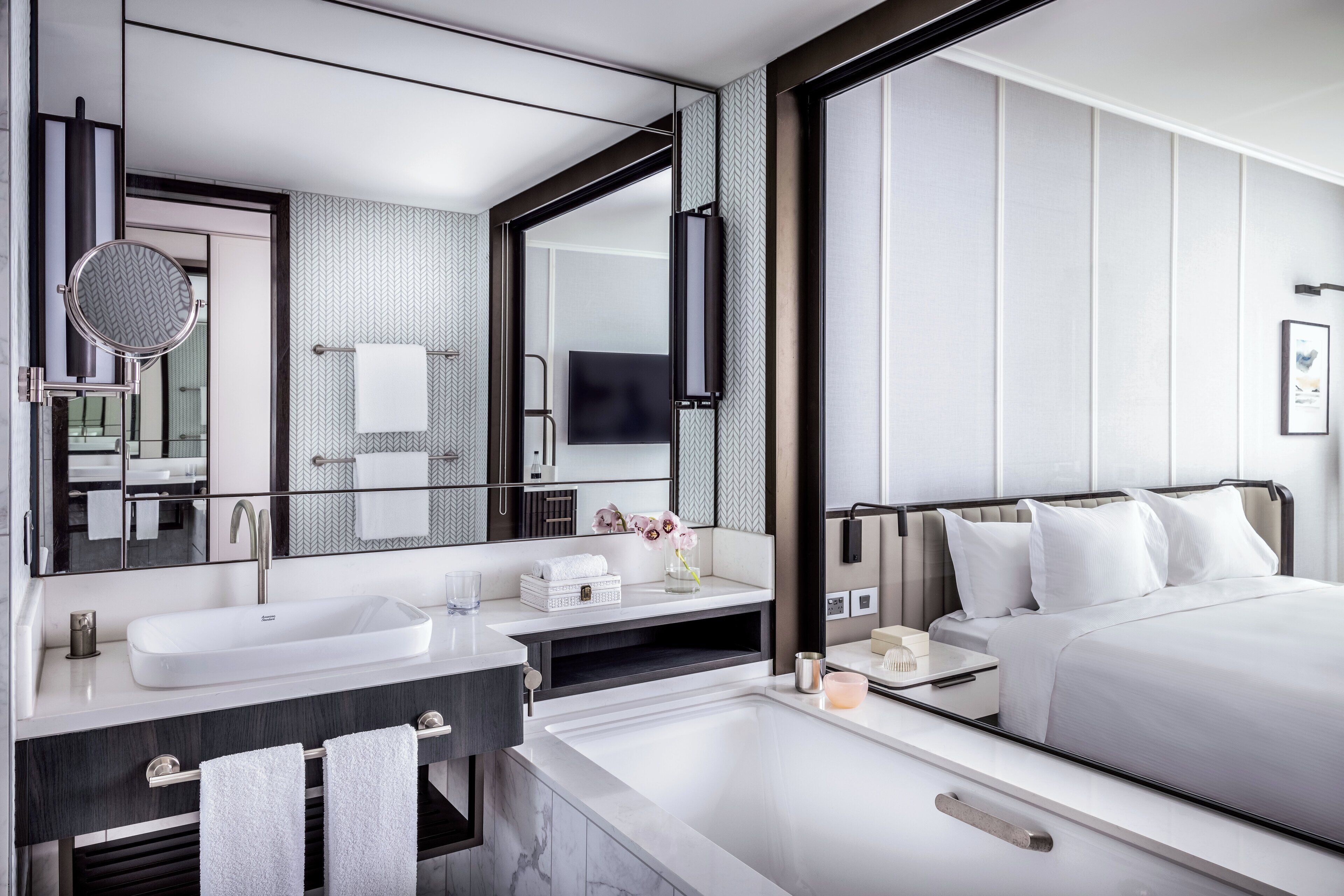 A modern hotel room with a large bed, a separate bathroom with a bathtub, a large mirror, and a flat-screen TV.