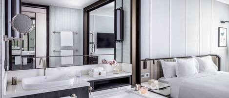 A modern hotel room with a large bed, a separate bathroom with a bathtub, a large mirror, and a flat-screen TV.