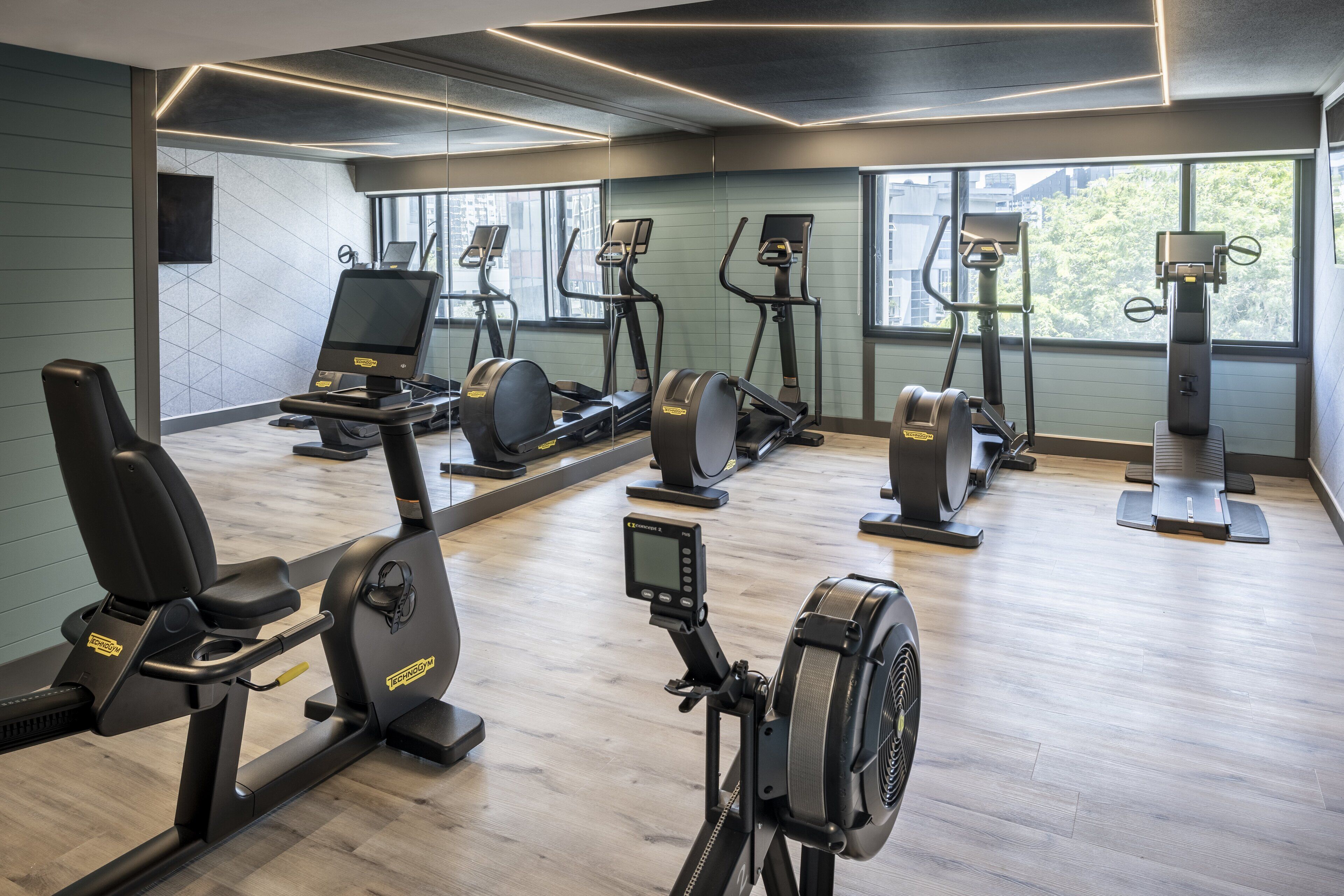 A modern gym with various cardio machines, a digital display, and large windows.