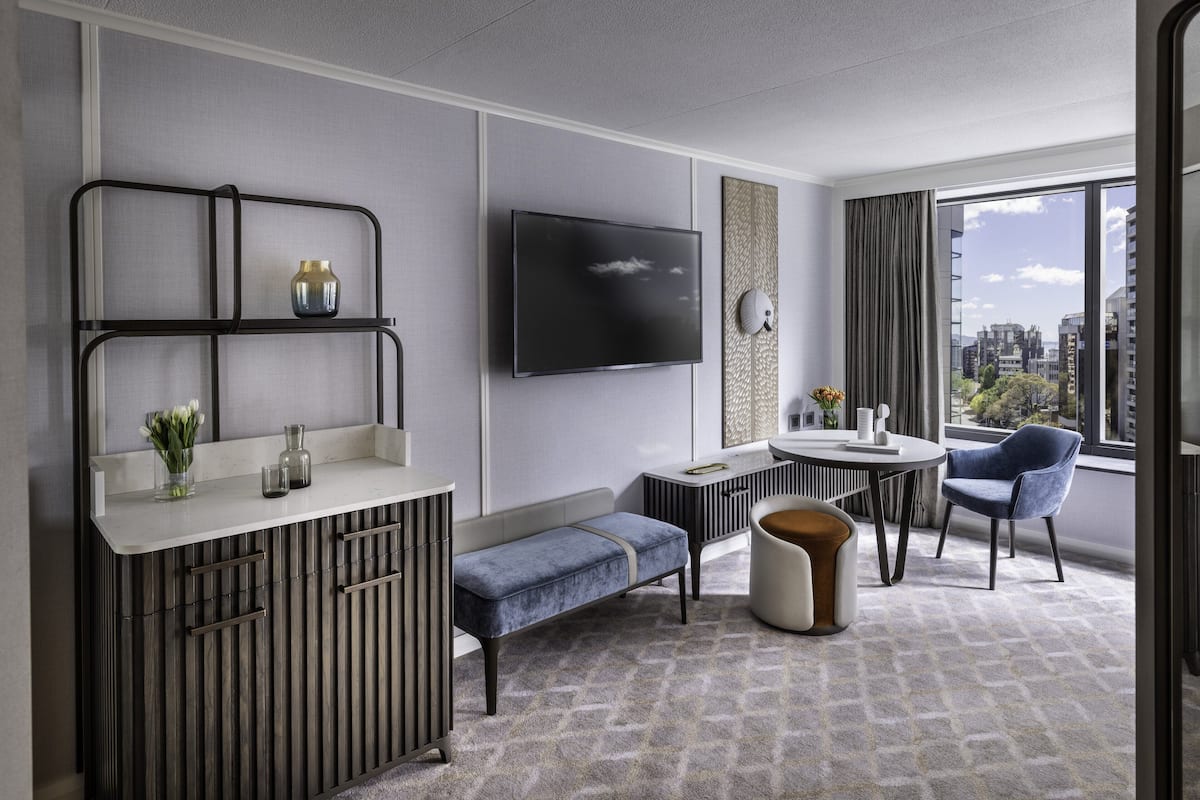 A modern hotel room with a flat-screen TV, a small dining table, a bench, and a view of the city through large windows.