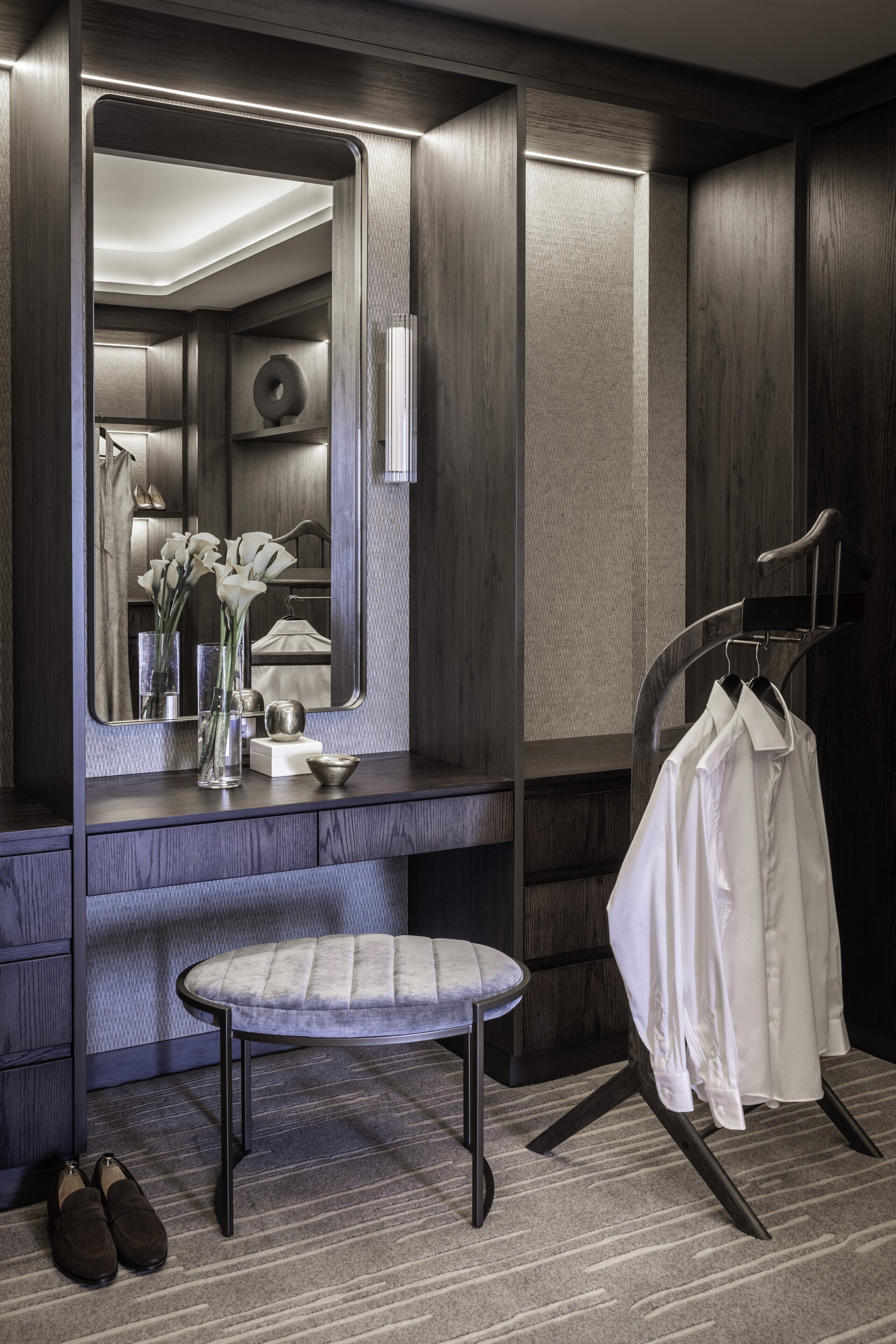 A modern bathroom with a large mirror, a vanity with a stool, a bench, and a hanging clothes rack.