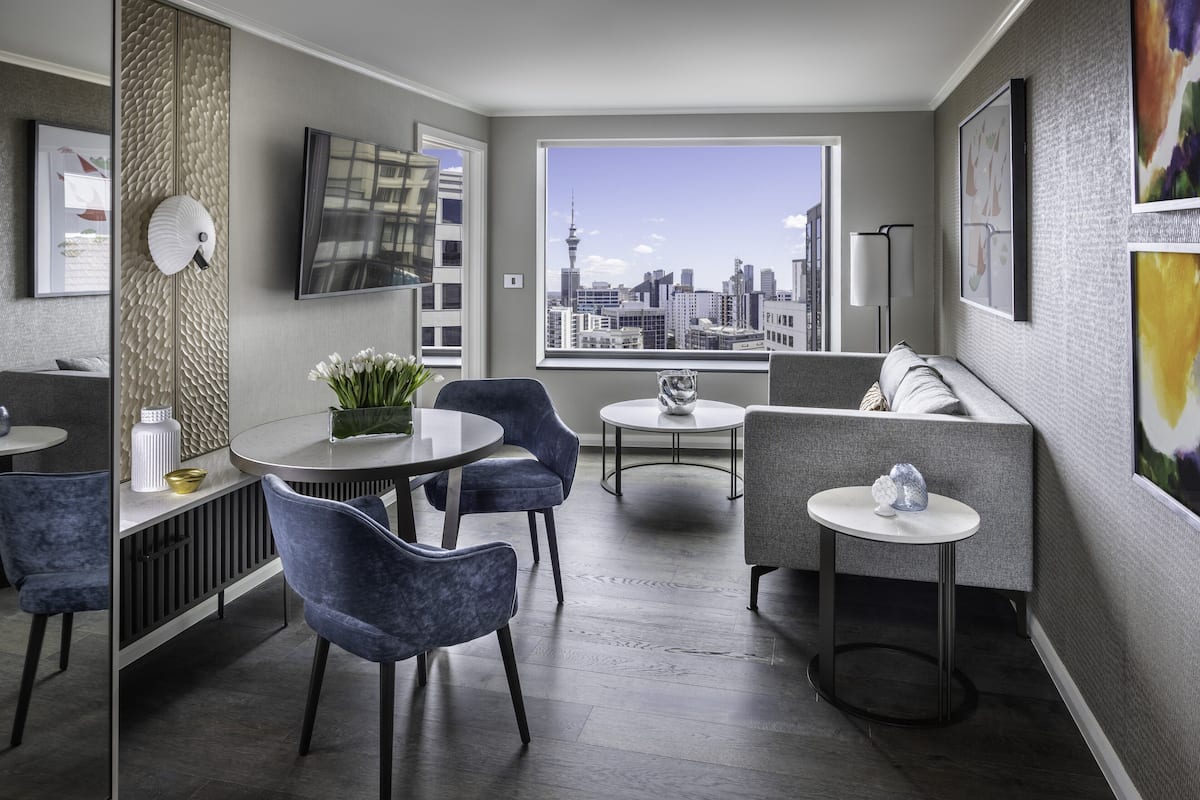 A modern hotel room with a city view, a round dining table, blue chairs, a sofa, and a small side table.