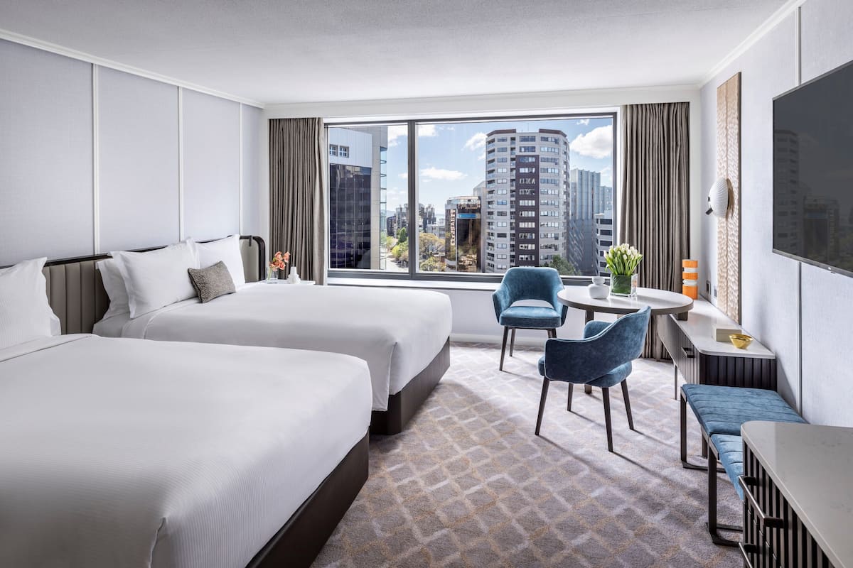 A hotel room with two beds, a desk, chairs, a TV, and a large window with a city view.