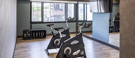 A gym with two stationary bikes, a TV mounted on the wall, and a window with a view of buildings outside.