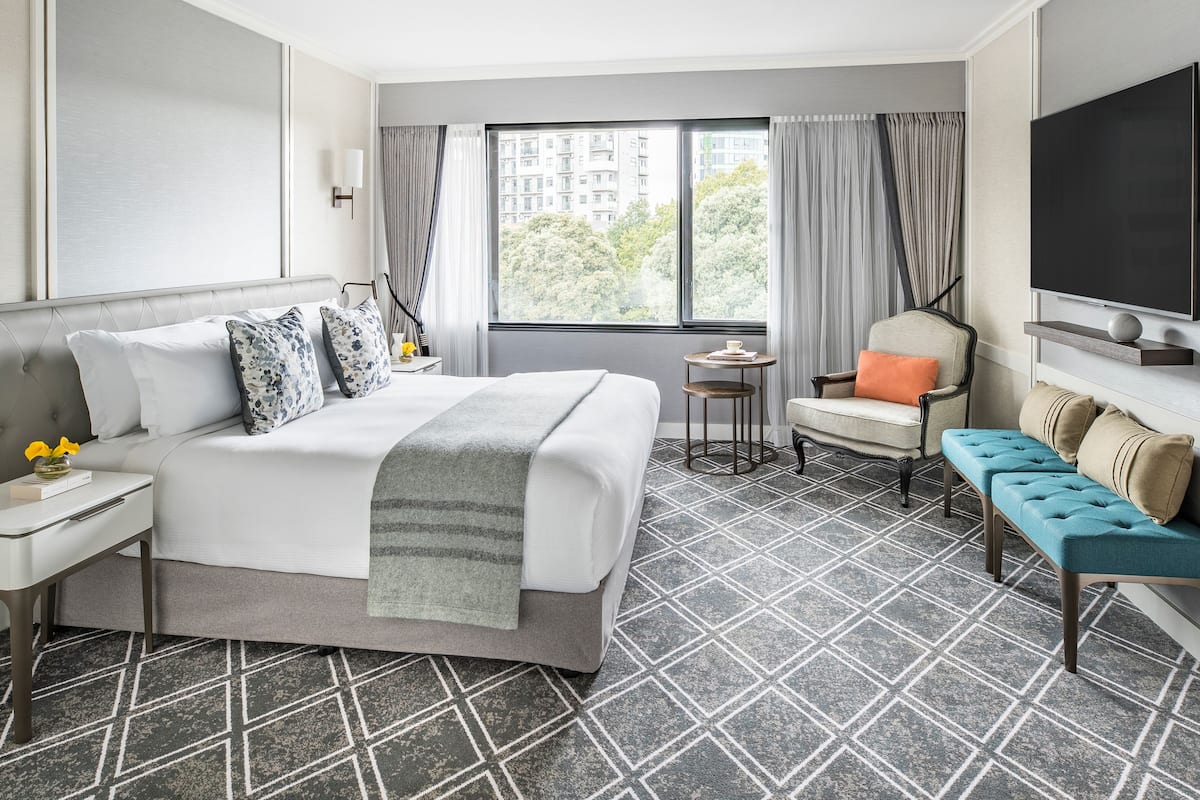 A hotel room with a large bed, a chair, a sofa, a TV, and a window with a view of buildings and trees.