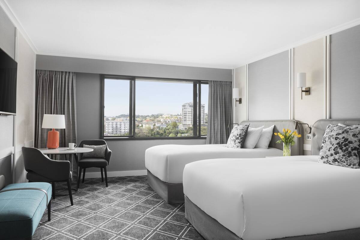 A hotel room with two beds, a desk, chairs, a lamp, and a view of the city.