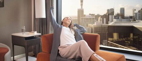 Una persona relajándose en un sofá con vista a un paisaje urbano, incluyendo una torre destacada, en una habitación moderna de hotel con una mesita auxiliar y una lámpara.