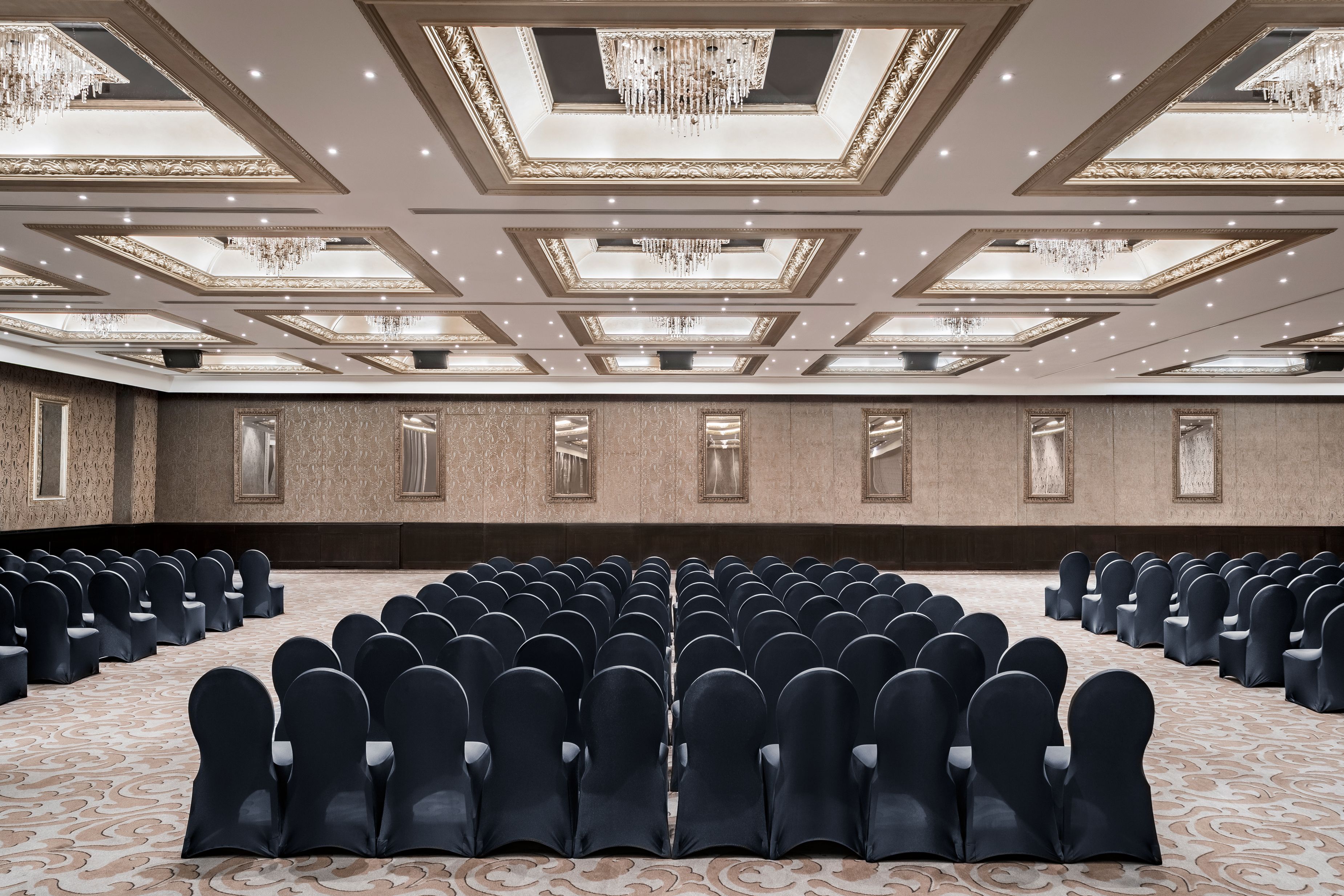 Una grande sala conferenze con file di sedie, un tappeto decorato e dettagli ornamentali al soffitto.