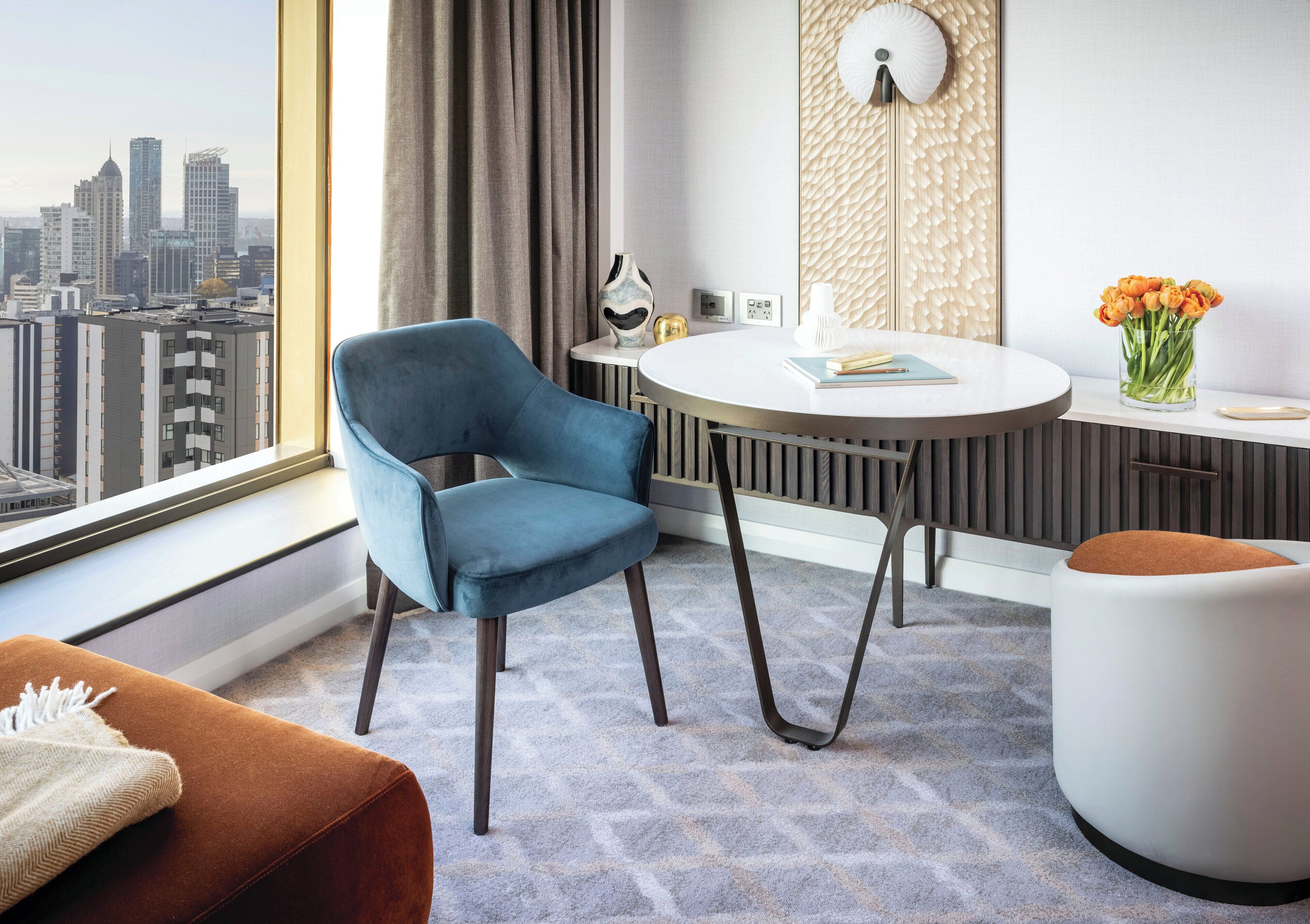 Une chambre d’hôtel moderne avec vue sur la ville, une table à manger ronde, un fauteuil bleu, un pouf blanc et un vase de fleurs.