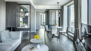 Een moderne hotelkamer met een grijze bank, een ronde salontafel, een eethoek met een witte tafel en stoelen, en een groot raam met uitzicht over de stad.