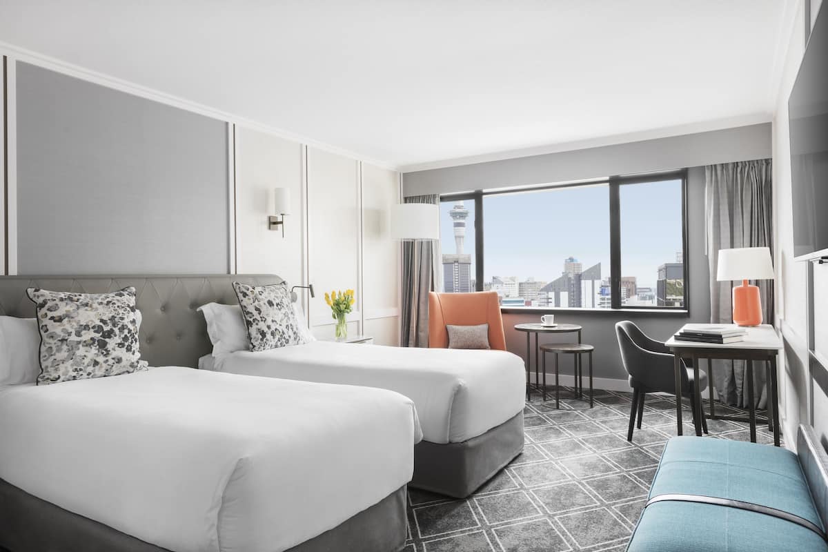 A hotel room with two beds, a desk, a chair, and a view of the cityscape.