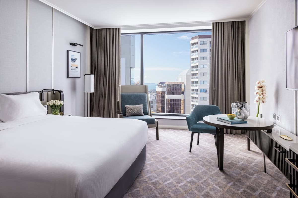 A hotel room with a large bed, a desk, a chair, a window with a city view, and a vase with flowers.
