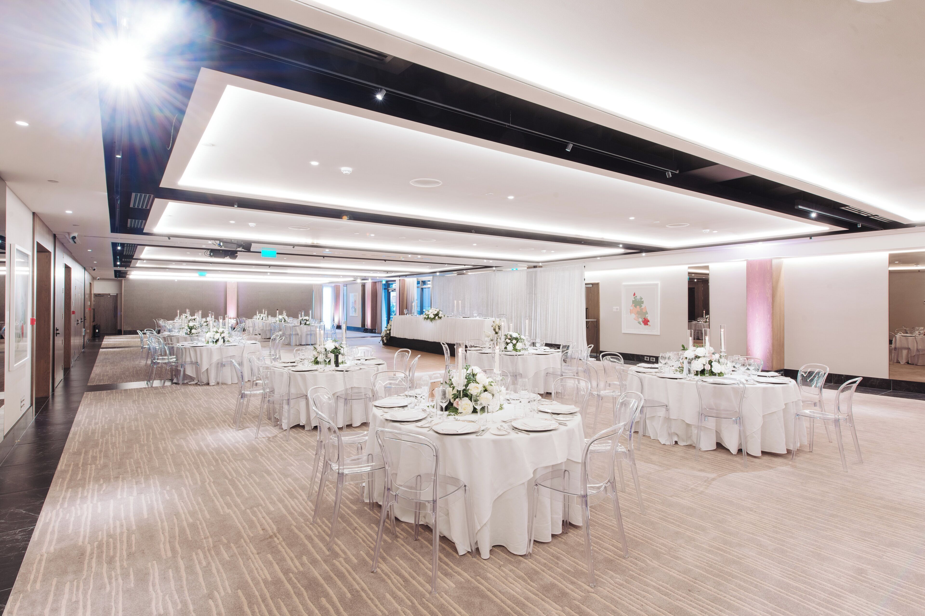 A spacious banquet hall with multiple round tables set for events, featuring clear chairs and white tablecloths.
