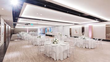 A spacious banquet hall with multiple round tables set for events, featuring clear chairs and white tablecloths.