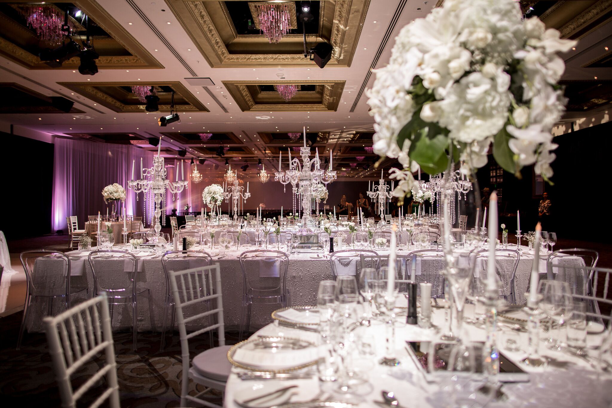 A banquet hall with round tables set for a formal event, featuring chandeliers and floral centerpieces.