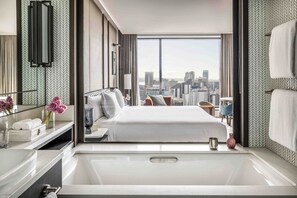 A modern hotel room with a large bed, a bathroom with a bathtub, and a city view through large windows.