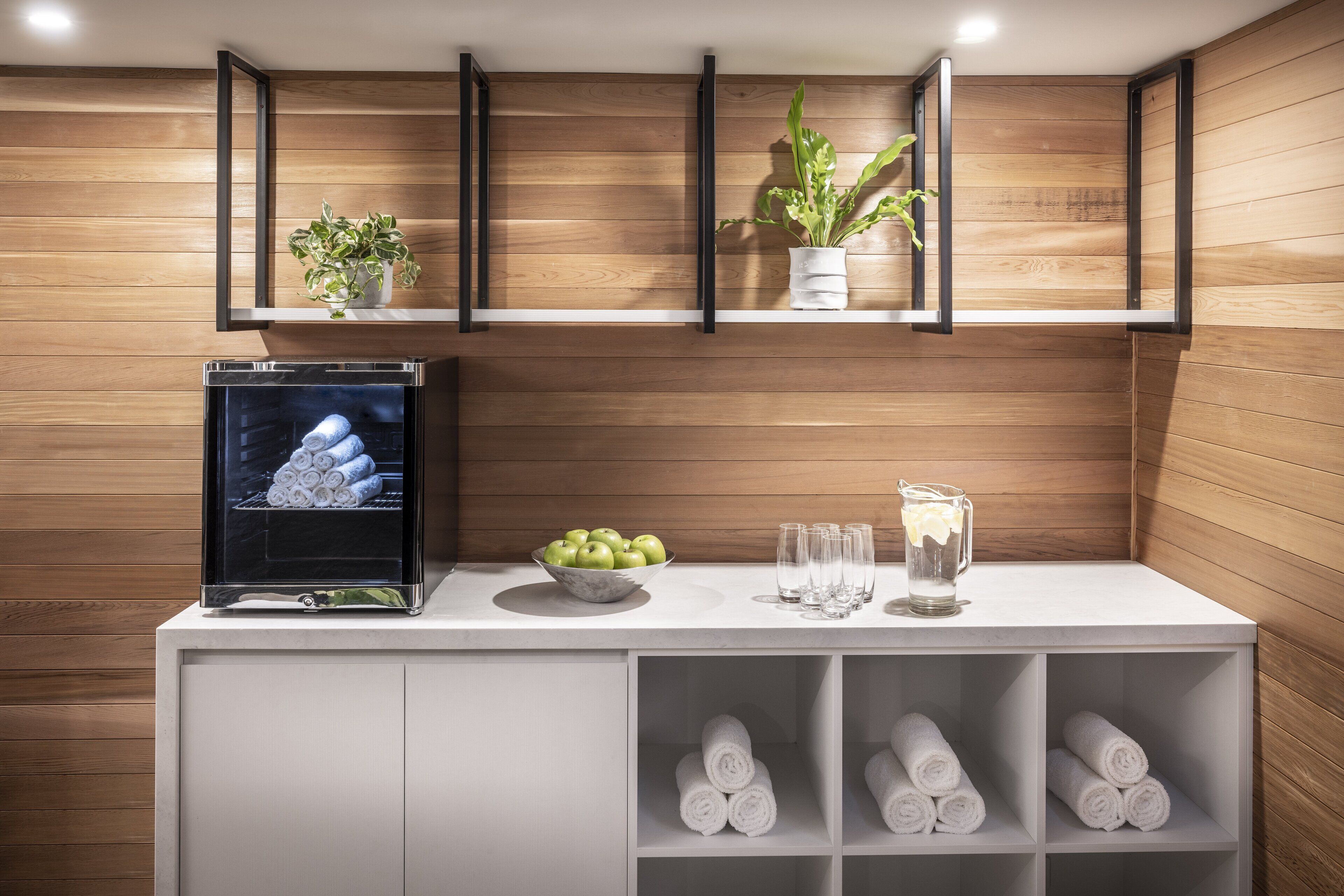 A modern bathroom with wooden paneling, a built-in TV, a sink with a bowl of apples, glasses, and a pitcher.