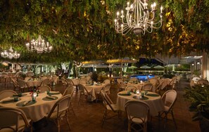 Un área de comedor al aire libre con mesas redondas y sillas de mimbre, rodeada de abundante vegetación e iluminación colgante.