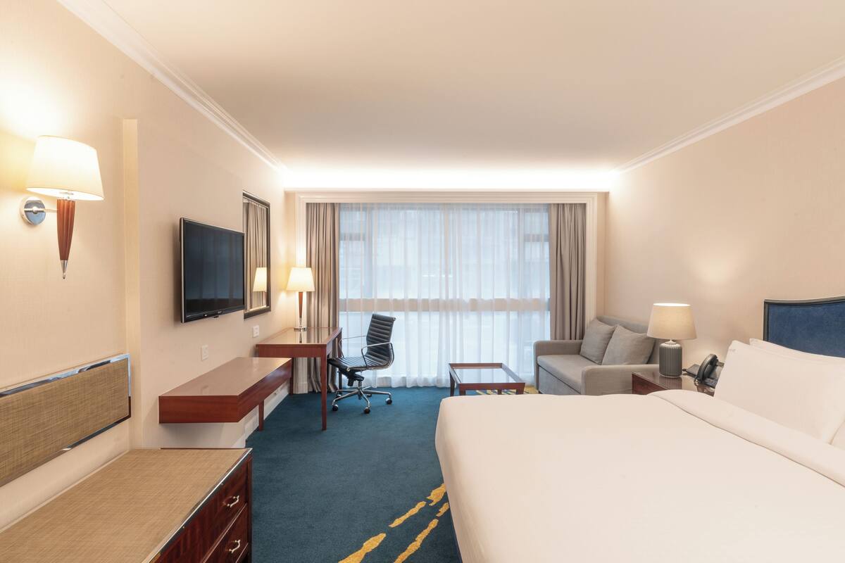 A hotel room with a large bed, a desk with a chair, a sofa, a television, and a window with curtains.